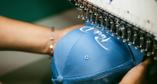 Hands positioning blue cap under embroidery machine needles